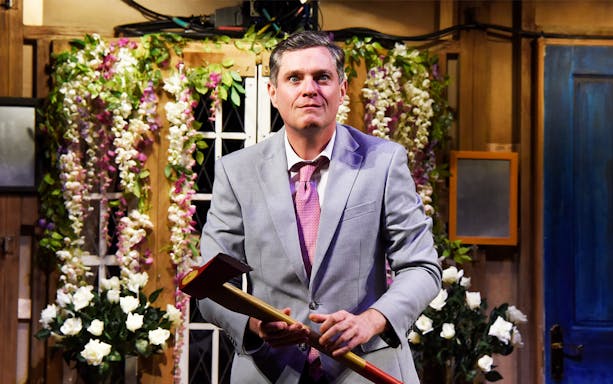 Man in a suit holding an axe on stage with floral backdrop, "Noises Off" play.