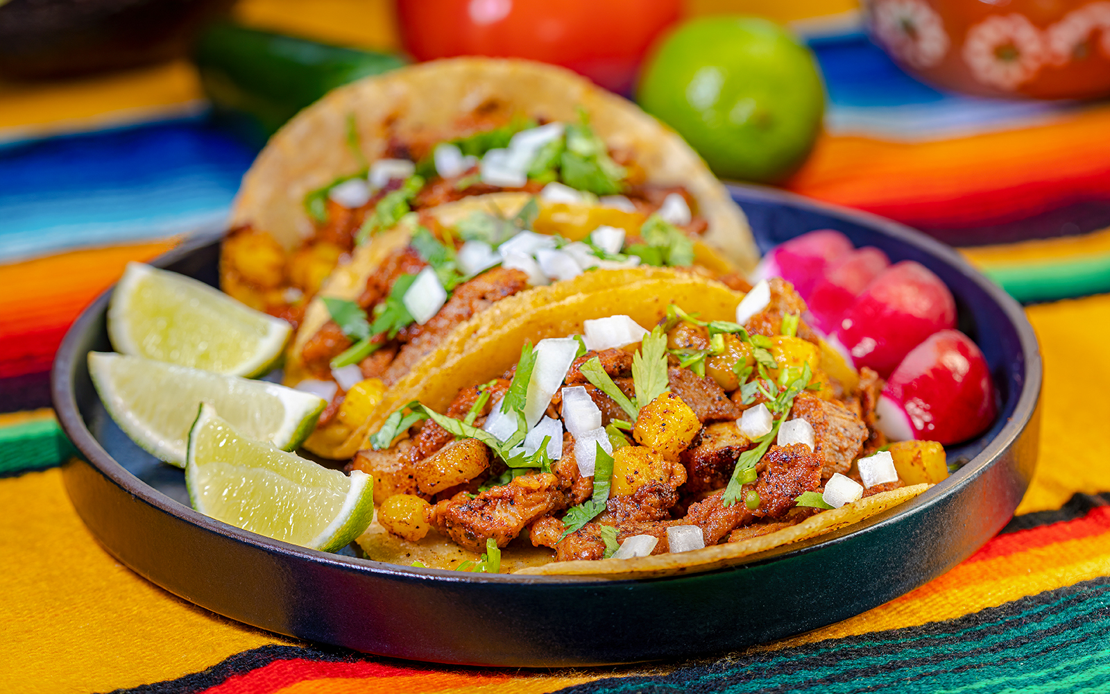 Al Pastor tacos with pineapple, onions, and cilantro on a colorful tablecloth.