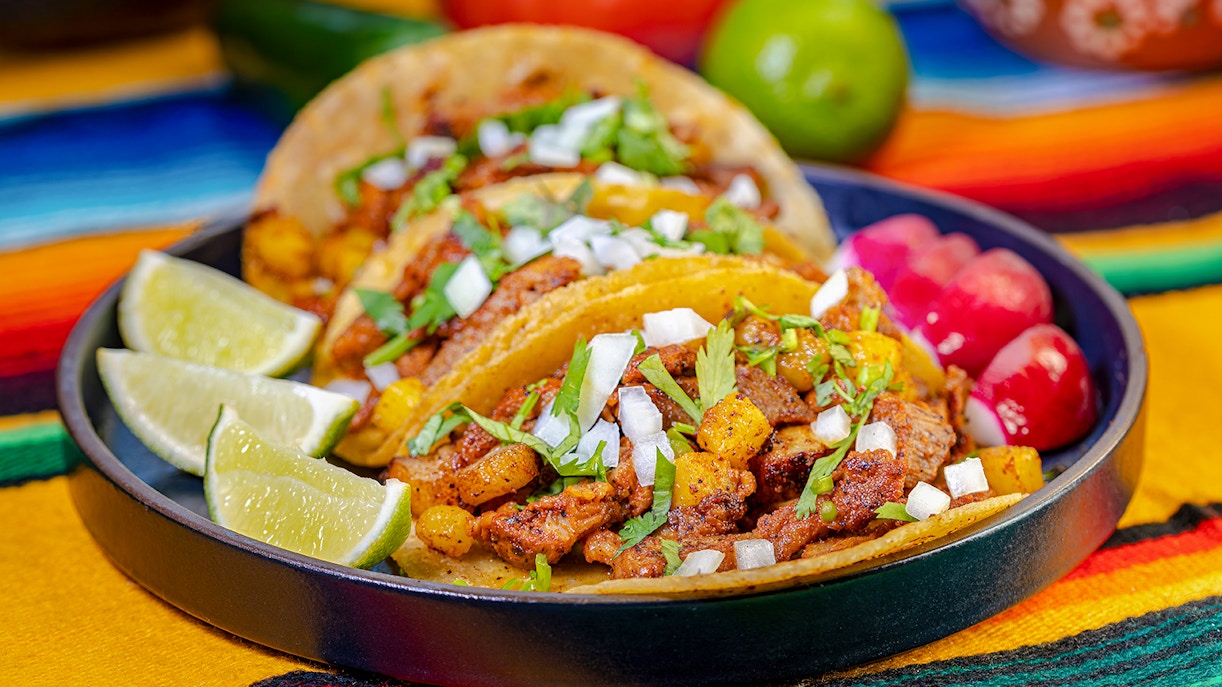 Al Pastor tacos with pineapple, onions, and cilantro on a colorful tablecloth.
