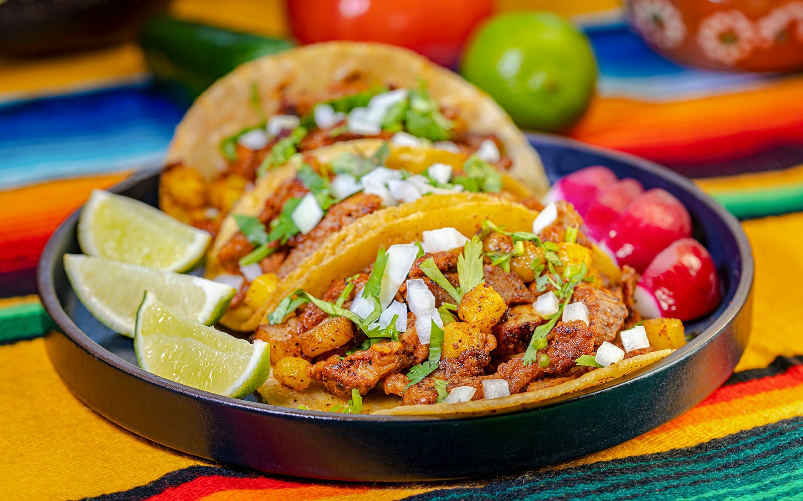Al Pastor tacos with pineapple, onions, and cilantro on a colorful tablecloth.