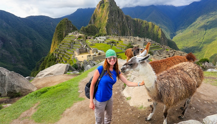 Traveler with llamas at Machu Picchu, Peru, with ancient ruins and mountains in the background.