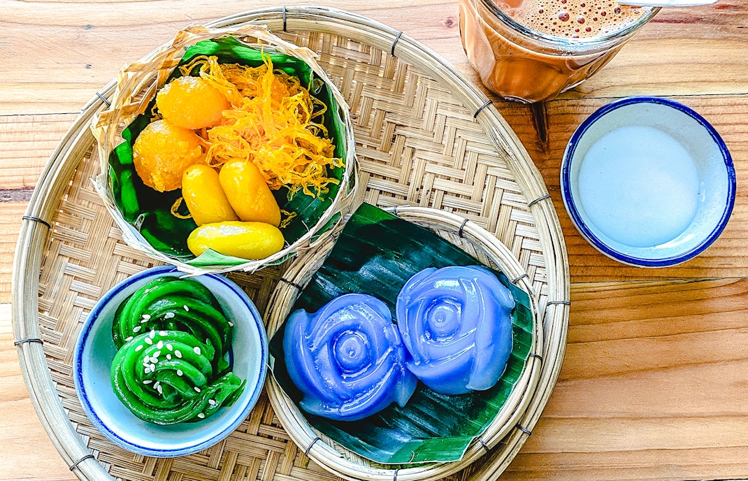Assorted traditional Thai desserts on a woven tray with tea and coconut milk.
