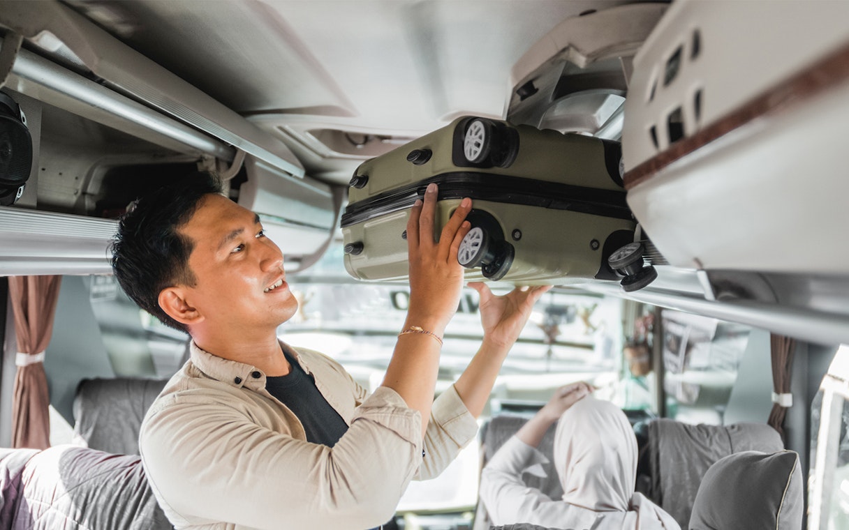 Man placing suitcase in overhead compartment on a bus.