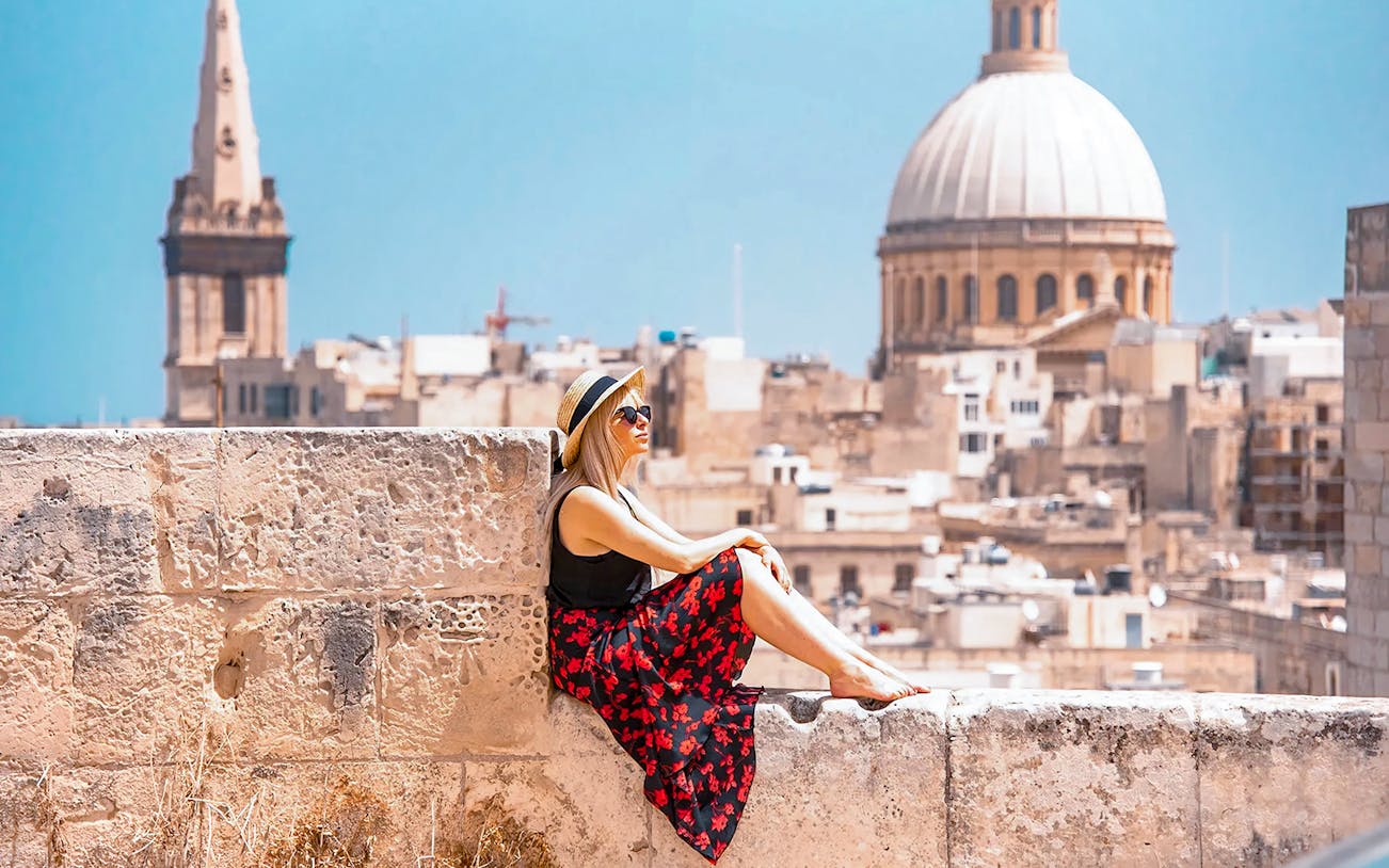 Woman sitting on a stone wall overlooking Valletta skyline with St. Paul's Cathedral in the background.