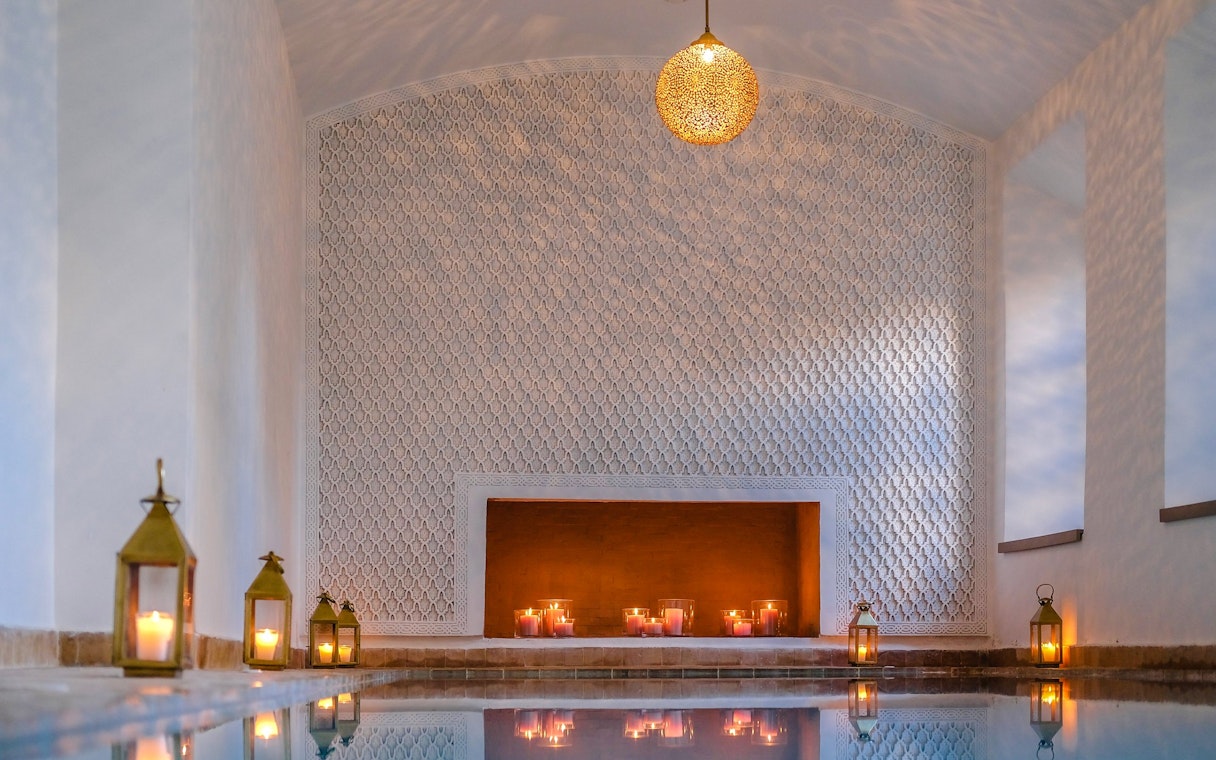 Luxurious steam hammam with ornate walls, candles, and lanterns reflecting on water.
