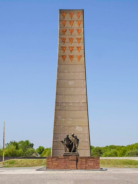 Sachsenhausen Memorial obelisk with statues, surrounded by greenery under a clear sky.