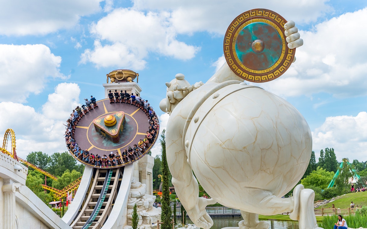 Visitors on a spinning ride at Astérix Park, France, with a large statue in the foreground.