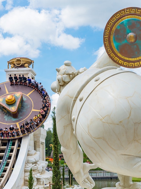 Visitors on a spinning ride at Astérix Park, France, with a large statue in the foreground.