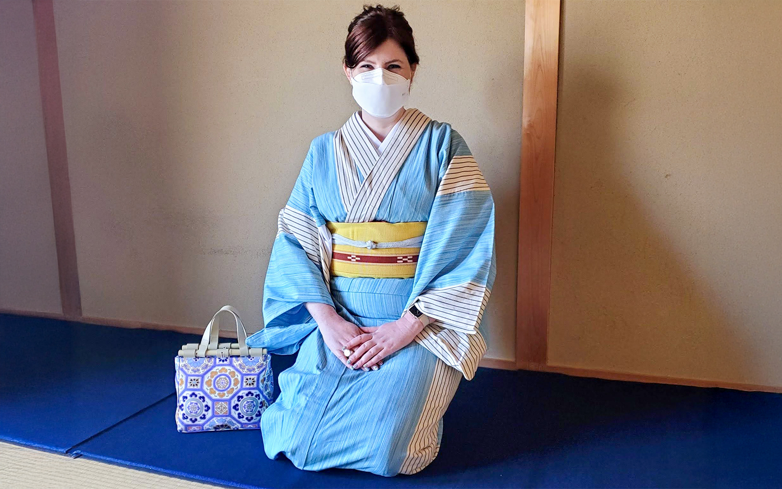 Woman in blue kimono and face mask kneels on tatami mat with decorative bag.