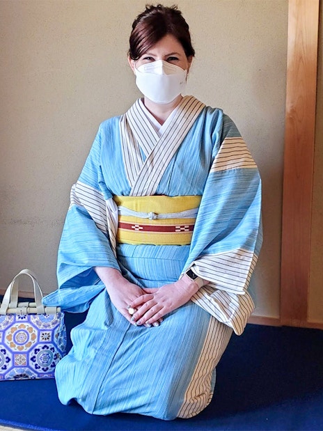 Woman in blue kimono and face mask kneels on tatami mat with decorative bag.