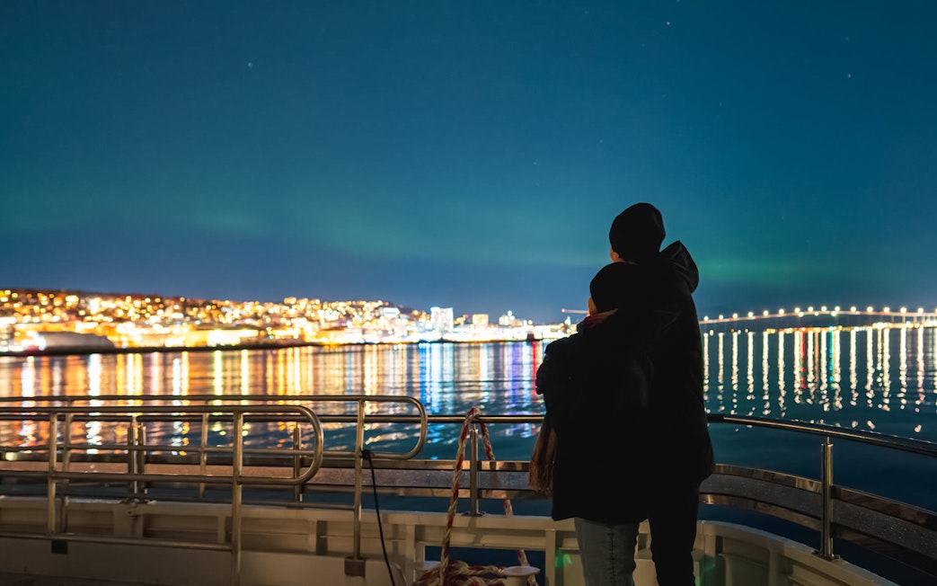 Couple on a cruise ship viewing Northern Lights over a lit cityscape.