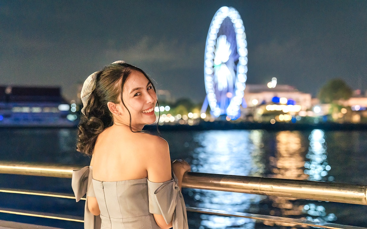 Female tourist enjoying night view on White Orchid River Cruise with Ferris wheel in background.