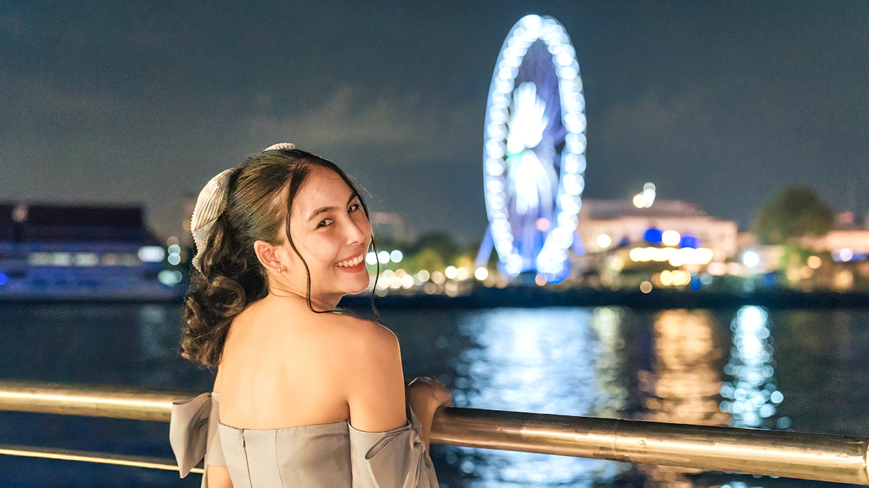 Female tourist enjoying night view on White Orchid River Cruise with Ferris wheel in background.