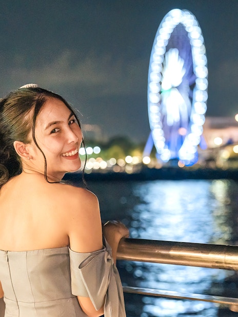 Female tourist enjoying night view on White Orchid River Cruise with Ferris wheel in background.