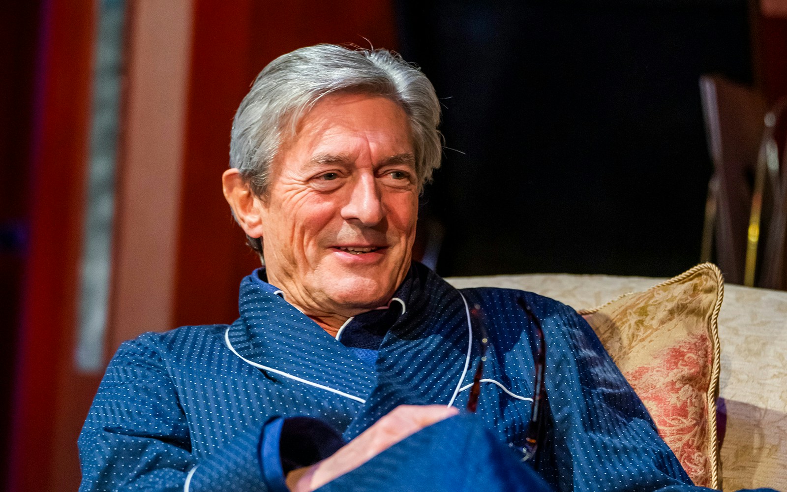 Man in blue robe smiling on a couch during "Private Lives" performance.