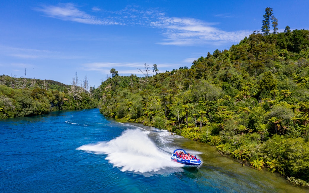 Jet boat speeding on Huka Falls river with lush green mountains in the background.