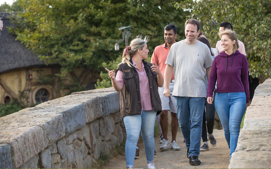 Tour guide leading group across stone bridge at Hobbiton Movie Set, surrounded by greenery.