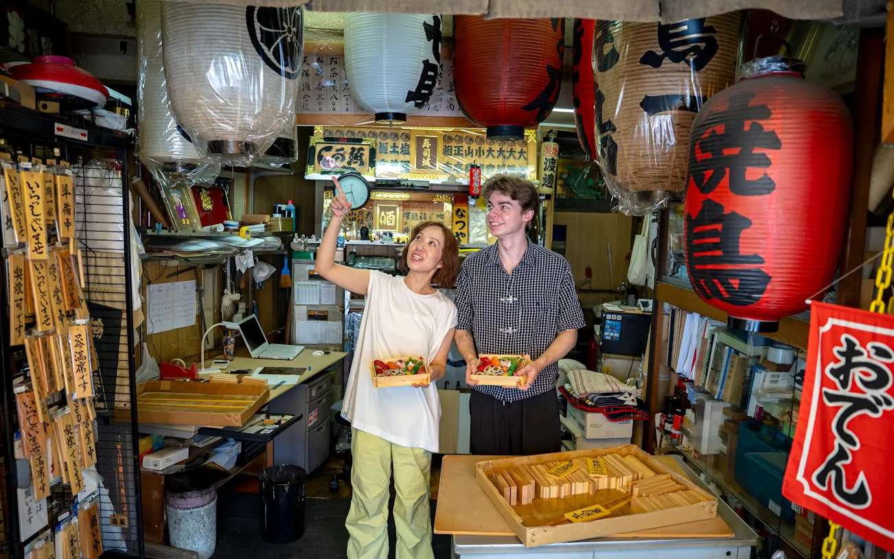Two people holding sushi bento boxes inside a Tsukiji Market shop with lanterns.