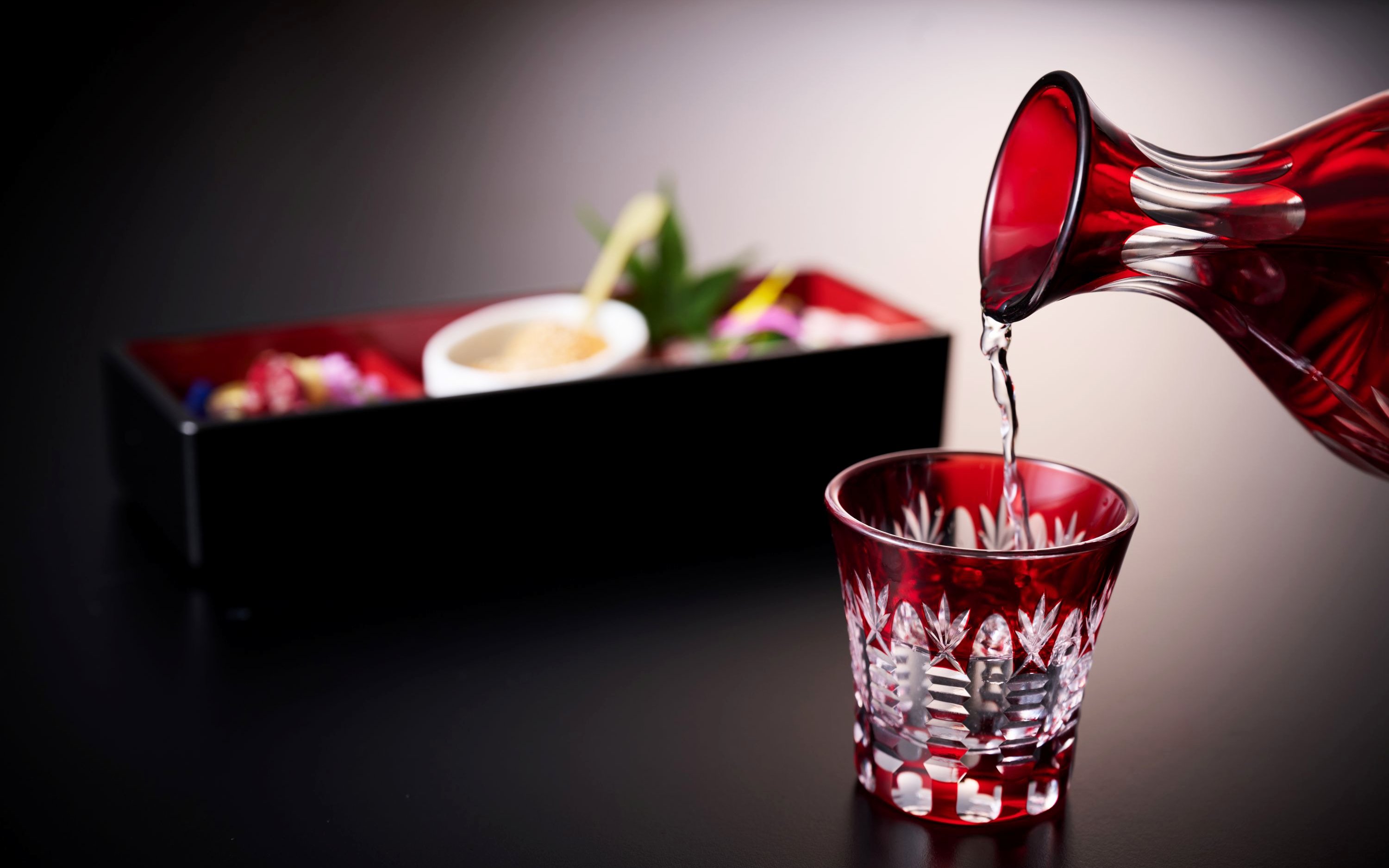 Pouring sake into a red glass with a traditional Japanese meal in the background.