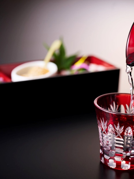 Pouring sake into a red glass with a traditional Japanese meal in the background.