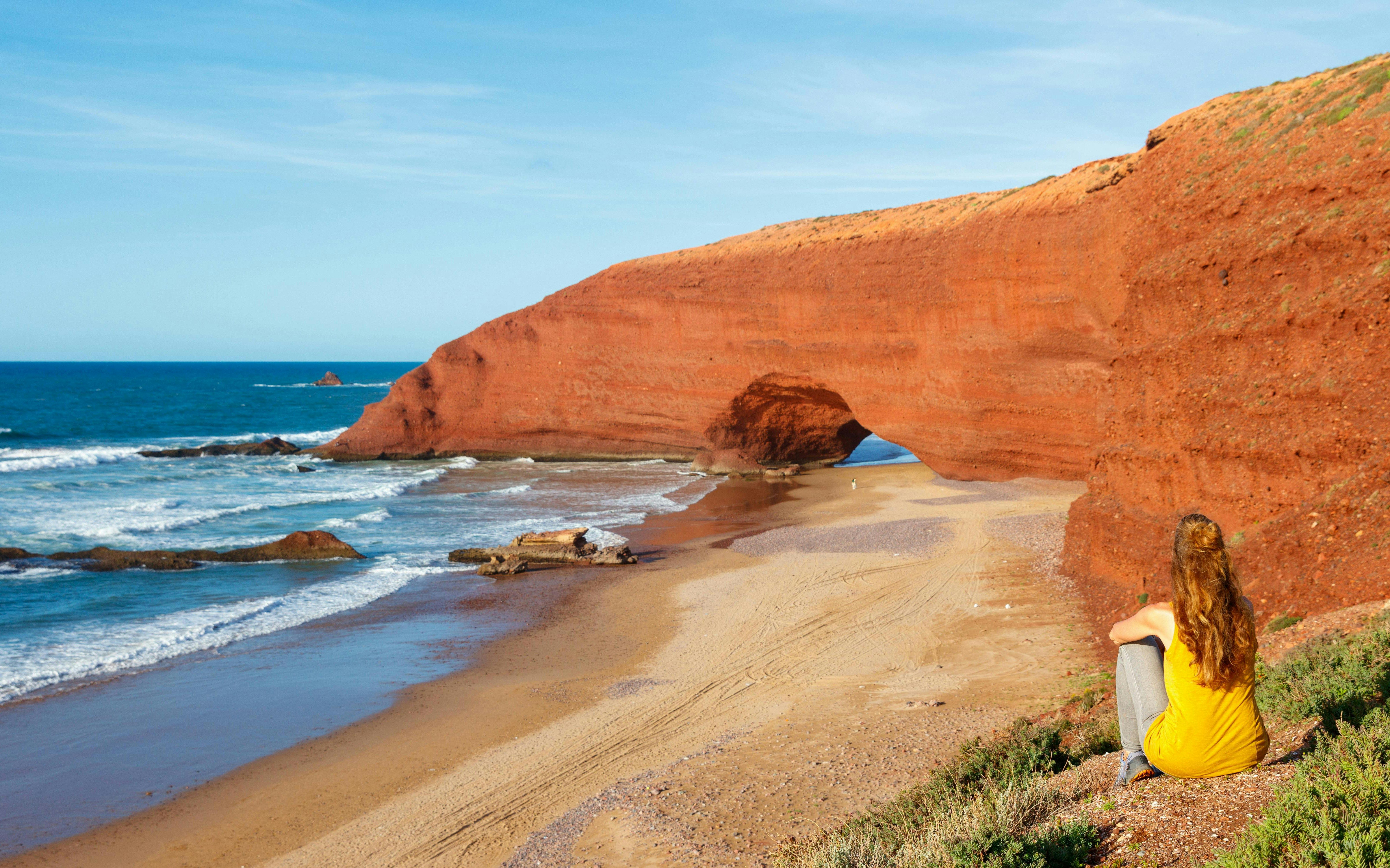 Woman sitting and viewing red rock arches at Legzira Beach, Morocco.