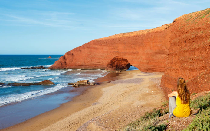 Woman sitting and viewing red rock arches at Legzira Beach, Morocco.