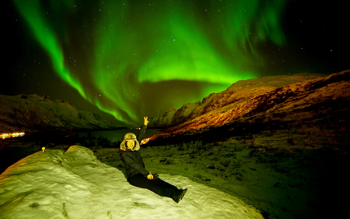 Tourist sitting on snowy hill under Northern Lights in Tromso, Norway.