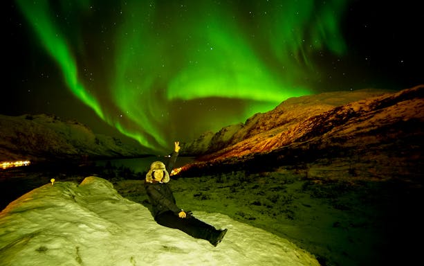 Tourist sitting on snowy hill under Northern Lights in Tromso, Norway.