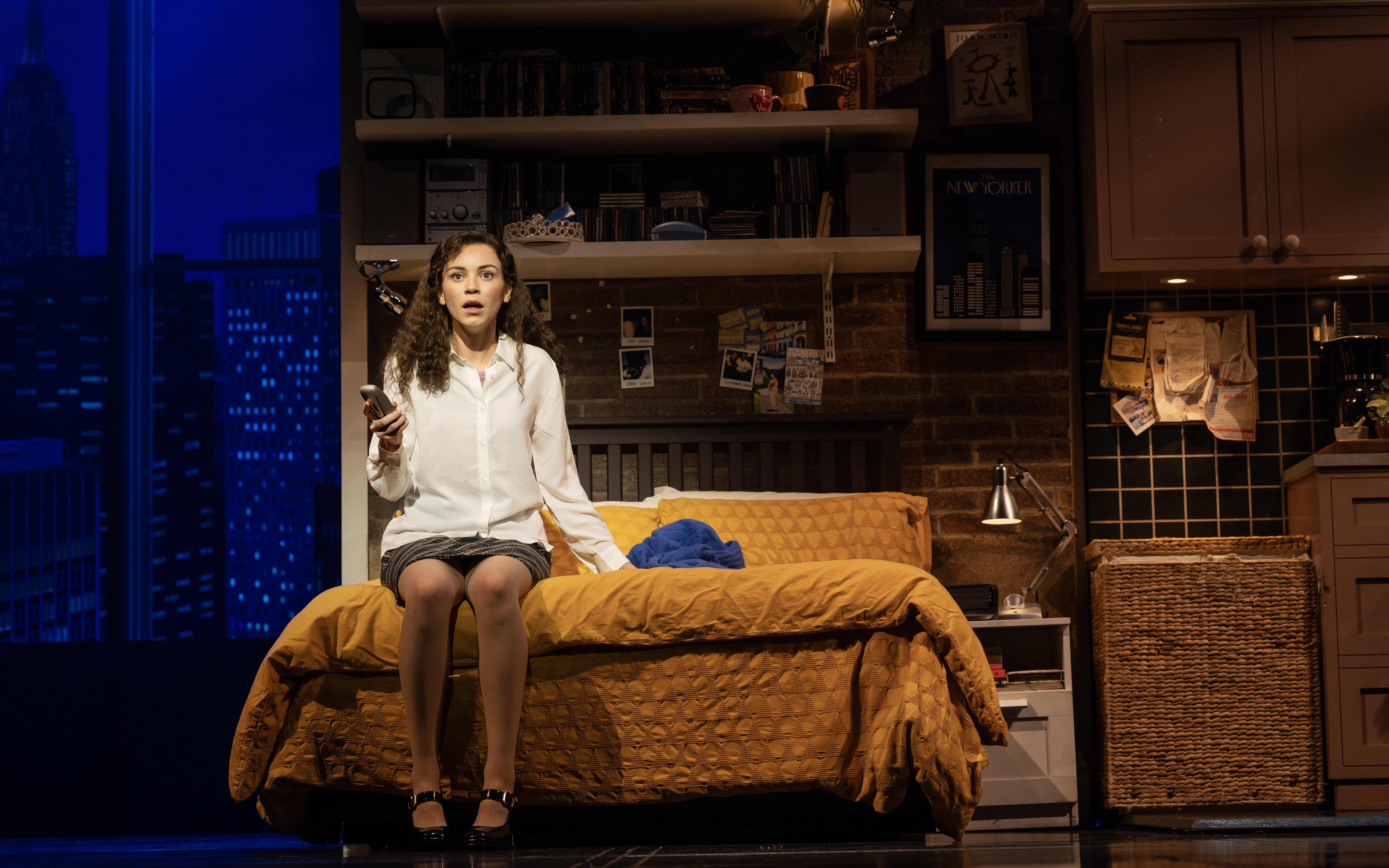 Stage scene from The Devil Wears Prada West End show with a woman sitting on a bed holding a phone.