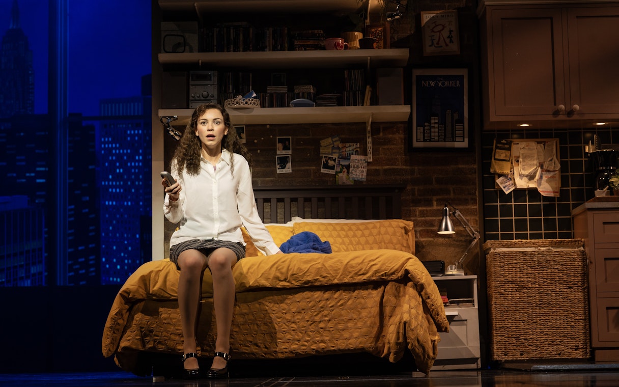 Stage scene from The Devil Wears Prada West End show with a woman sitting on a bed holding a phone.