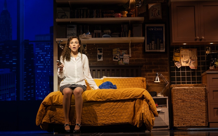 Stage scene from The Devil Wears Prada West End show with a woman sitting on a bed holding a phone.