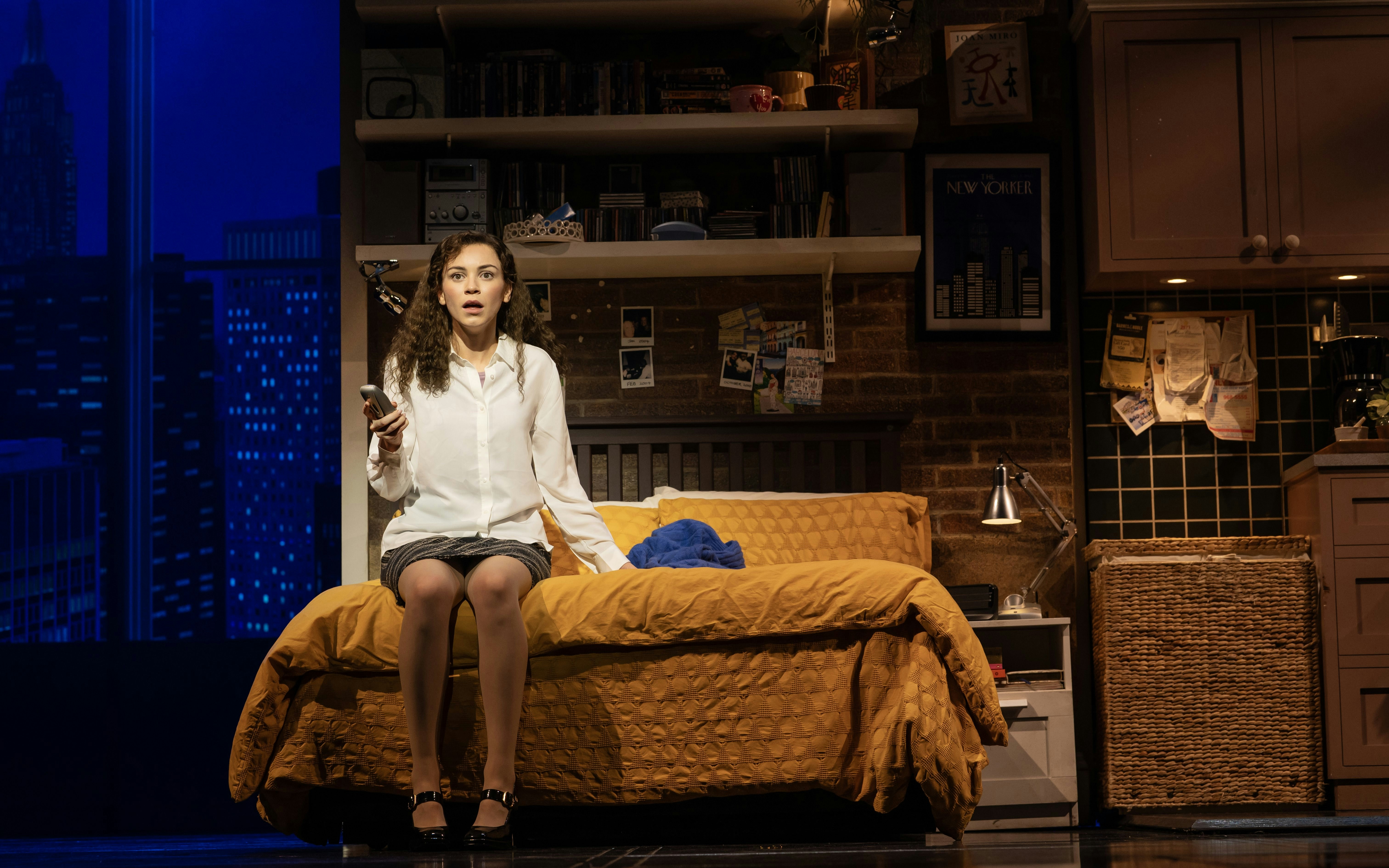 Stage scene from The Devil Wears Prada West End show with a woman sitting on a bed holding a phone.
