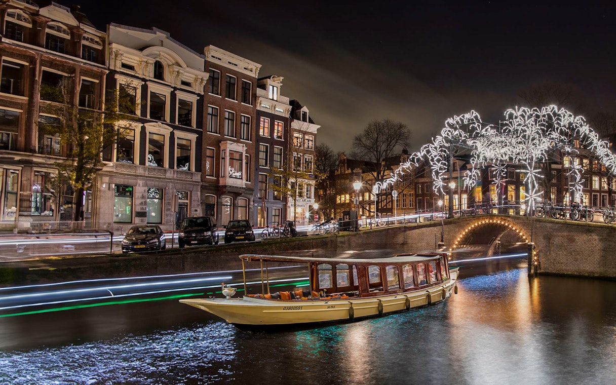 Canal boat cruising under illuminated bridge during Amsterdam Light Festival.