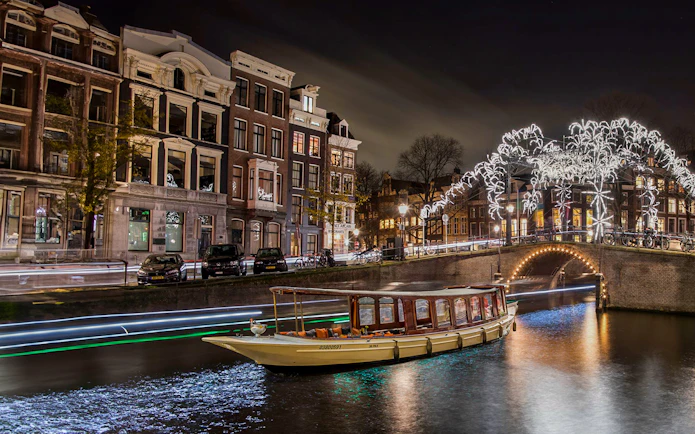 Canal boat cruising under illuminated bridge during Amsterdam Light Festival.