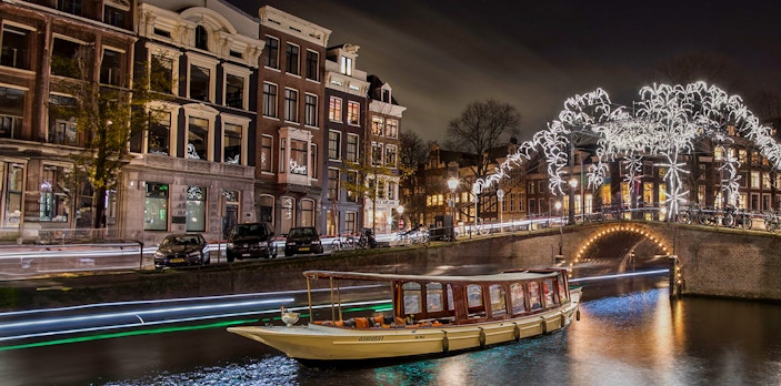 Canal boat cruising under illuminated bridge during Amsterdam Light Festival.