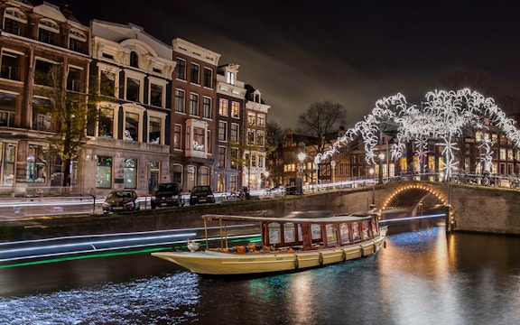 Canal boat cruising under illuminated bridge during Amsterdam Light Festival.