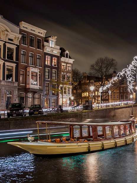 Canal boat cruising under illuminated bridge during Amsterdam Light Festival.