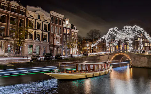 Canal boat cruising under illuminated bridge during Amsterdam Light Festival.