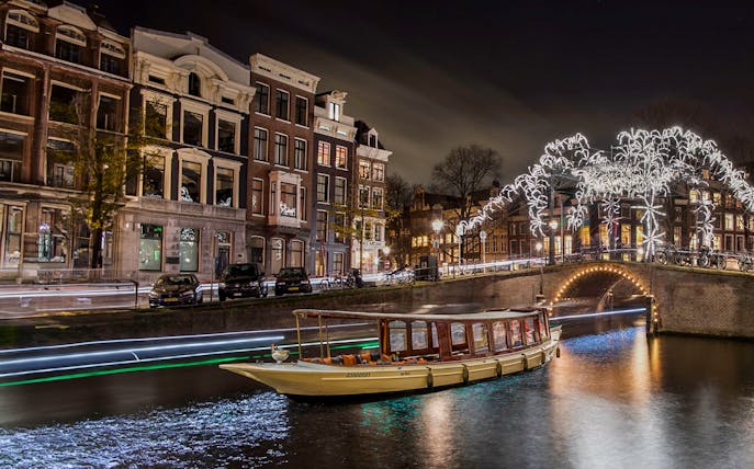 Canal boat cruising under illuminated bridge during Amsterdam Light Festival.
