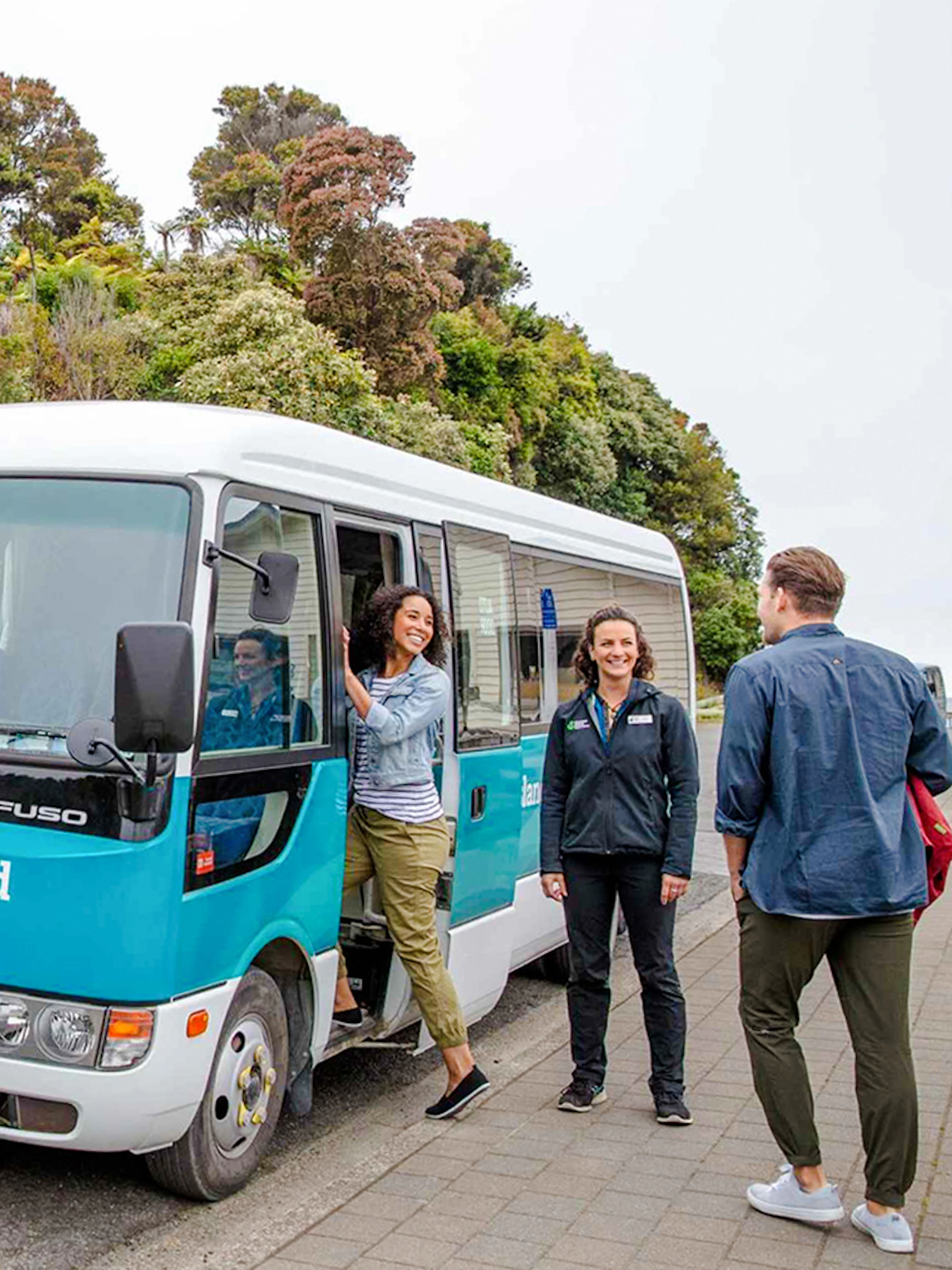 Stewart Island tour bus parked near a red building with people boarding and interacting.