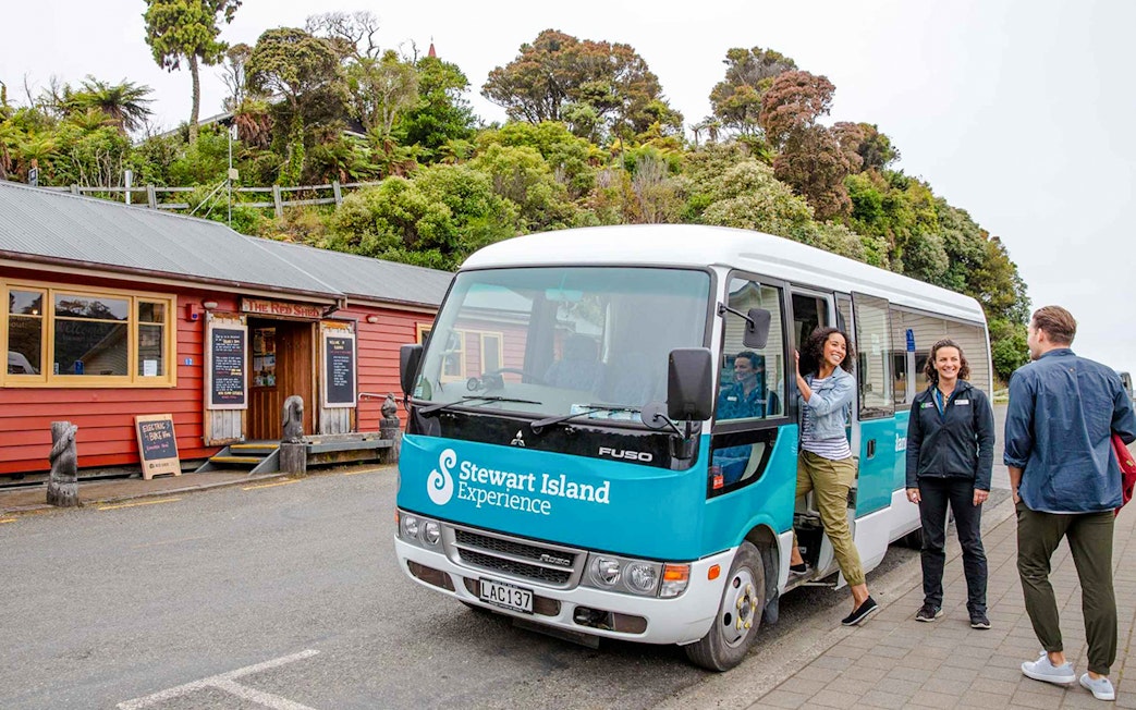 Stewart Island tour bus parked near a red building with people boarding and interacting.