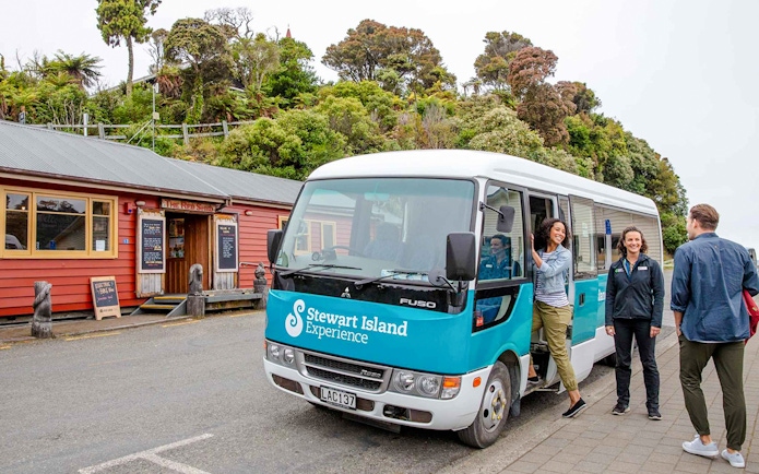 Stewart Island tour bus parked near a red building with people boarding and interacting.