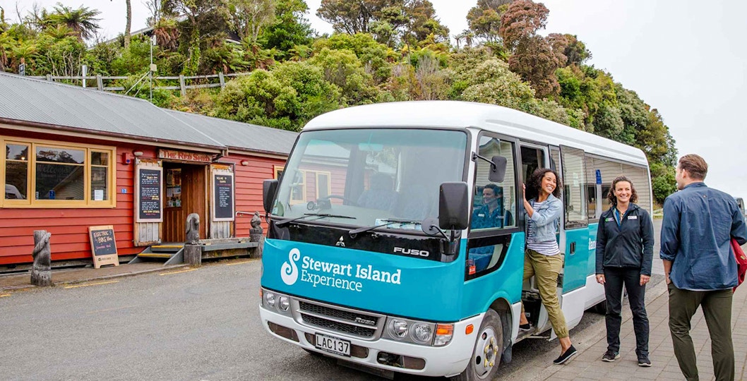 Stewart Island tour bus parked near a red building with people boarding and interacting.