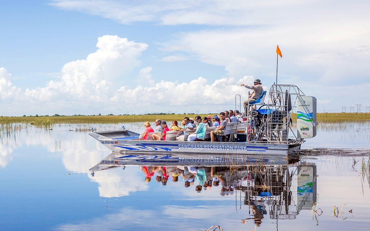 Airboat with tourists gliding through Everglades wetlands under a clear sky.