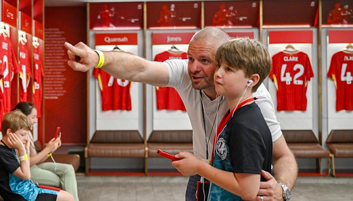 Father and son with audio guides in Liverpool stadium locker room.