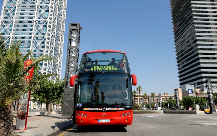 Red double-decker bus on Barcelona City Hop On Hop Off Tour near modern buildings.
