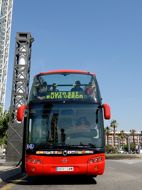 Red double-decker bus on Barcelona City Hop On Hop Off Tour near modern buildings.