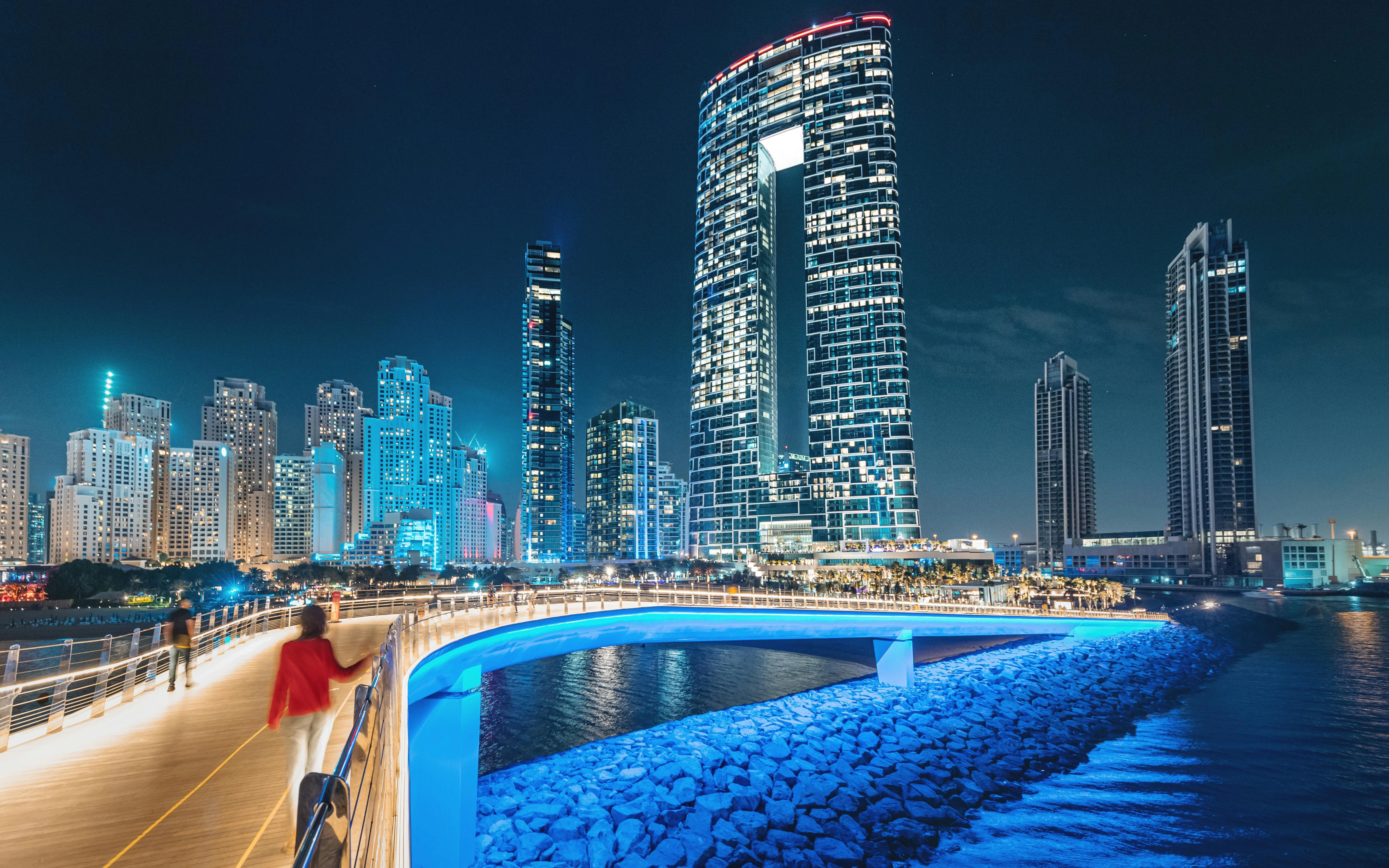 Footbridge with tourists leading to Dubai skyscrapers on Persian Gulf coast at night.