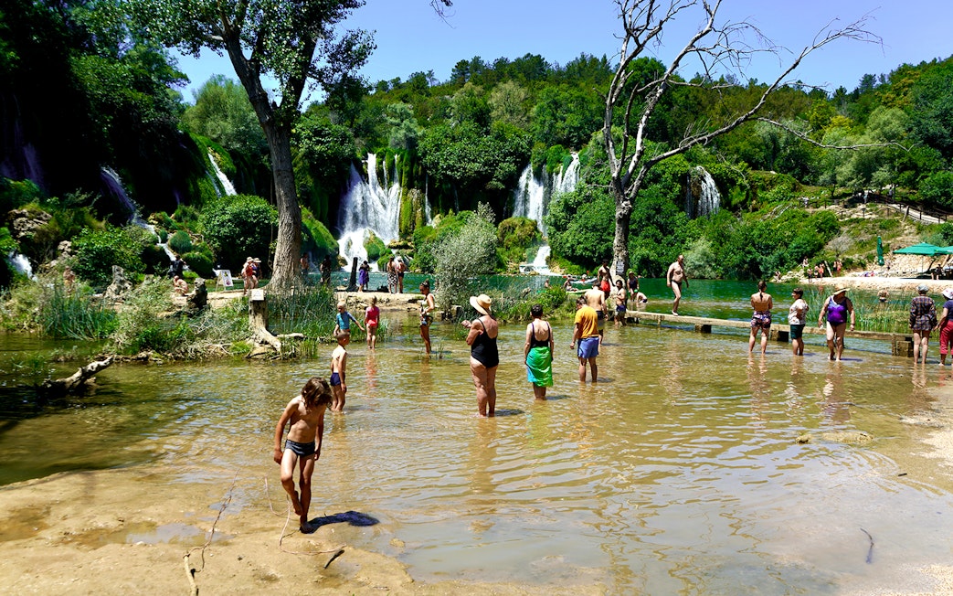 Tourists wading in water at Kravica Falls, Bosnia, with lush greenery and waterfalls in the background.
