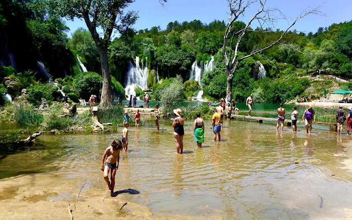 Tourists wading in water at Kravica Falls, Bosnia, with lush greenery and waterfalls in the background.